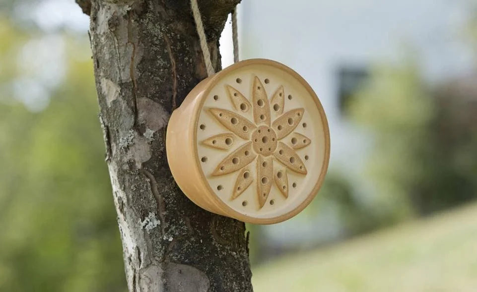 Handgefertigtes Wildbienenhotel aus sandsteinfarbener Keramik, aufgehängt an einem Baum – nachhaltige Nisthilfe für Bienen und Insekten.